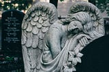 A stone statue of an angel leans peacefully against a graveyard headstone.