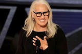 Jane Campion smiling at the podium, with her hand to her heart, as she gives her acceptance speech for Best Director at the 2022 Oscars.