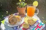 A table with a multi-colored cloth, a plate with Mexican biscuits on it, a flower pot, and a glass teacup with cinnamon tea in it.