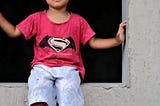 kid in a Superman shirt sits on a windowsill, balancing self by stretching out hands to touch the sides of the window frame