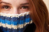 redheaded young woman has a turtleneck sweater pulled up over her mouth and nose, so that only her eyes are showing.