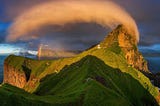 a rainbow peeks from under a billowing cloud arcs across a craggy mountain range