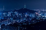 A night time photo of the city of Seoul Korea. In blue and black colours with lots of white lights.