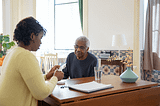 A woman in a yellow sweater preparing medicine for an elderly man in a blue sweater.