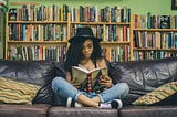 A young woman in a hat and blue jeans reading a book. She is sitting on a black, leather couch with her legs crossed. She is indoors, with a row of bookshelves behind her.