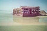A photograph using a long exposure of a concrete bunker on a beach. The water around it is blurred, suggesting a passage of time. There are graffiti painted on the bunker and the colours of the image have been muted, perhaps also suggesting the subject is from another era.