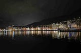 A nighttime waterfront view of Bryggen in Bergen, Norway, featuring a row of brightly lit, colorful wooden buildings with steep gabled roofs. Their reflections shimmer on the calm water in front. Behind the historic harbor district, a hillside dotted with lights from homes rises into the dark sky, adding depth to the scenic and culturally rich landscape.