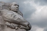 Profile photo of the Martin Luther King, Jr. Memorial in Washington, D.C., showing the leaders face and crossed arms carved in stone with a cloudy sky behind.