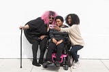 A non-binary person with a cane, a non-binary person sitting in a power wheelchair, and an invisibly disabled femme smiling and taking a cell phone selfie together.