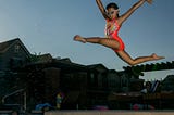 A little girl, wearing goggles and an orange swimsuit, leaps into a neighborhood pool.