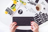 What You Need to Know Before You Start Blogging — Blogging Tips. Post by David Mania (www.davidmania.com). This image displays a woman typing on a black keyboard with posters and a coffee on the table beside the keyboard.