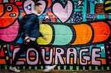 A man walks in front of a brightly painted wall. The word courage is written in block letters on the wall.