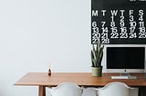 Wood work desk with a computer screen on it. Black calendar with bold white numbers on it is attached to the wall.