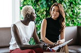 Two women sit next to one another referring to a piece of paper.