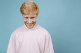 Image shows red-haired man in pink shirt against a blue background, grinning maniacally at camera.