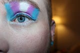 Close up photograph of half of a face of a caucasian youth, wearing studs earrings, and shorn dirty blond hair. The photo is cenbtered on their half of their face, one blue eye and eyebrow. Above the eyebrow is the trans flag in eyeshadow extending to the lower eyebrow.