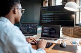 Photo of a Black person with glasses sitting in front of two laptops and a desktop computer, looking at code on the screens.
