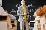 Students sat at their desks looking at a teacher standing in front of a blackbord