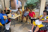 Six women sitting around a table outside an apartment building looking at the camera.