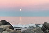 A sunset over crashing waves on a rocky shore. The sky is blue and pink. The sun is a yellow ball.