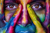 Close-up of a face with hands framing the eyes, covered in vivid, multicolored paint. The eyes are prominently visible, surrounded by bold hues of red, yellow, green, blue, and purple.