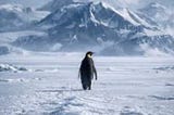 A solitary penguin walking across a vast snowy landscape toward distant mountains,