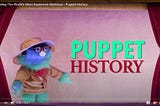 A blue puppet in a beige jacket with a red bowtie stands in a red and gold puppet theatre alongside the Puppet History Logo