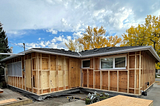 Image of a house from the outside with no siding, with framing extending past the original walls and window frames.