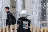 Person with a shiny disco ball as a head sitting on a bench reading an ‘ideas’ magazine while someone who looks aloof walks past in the background.