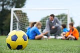 A football on a pitch with a coach and player out of focus in the background