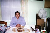 The photo shows the author’s father on his birthday. He’s a man with brown skin wearing a lilac shirt and is sat down at a table smiling in front of a chocolate birthday cake.