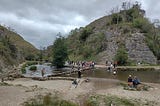 A picturesque scene from the Peak District. A river runs between two rocky hills. A small crowd of people is about to cross the river on stepping stones. Others are paddling in the river or sat by its banks.