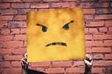 Two hands hold up a square sign with an angry face on it in front of a painted red brick wall.