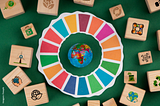 Vista superior de um globo terrestre no centro de um círculo colorido da Agenda 2030, cercado por blocos de madeira com ícones de sustentabilidade sobre fundo verde.