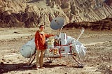 Carl Sagan stands beside a lunar lander craft in an orange jacket and beige slacks.