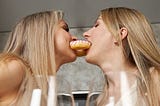 Two blonde women gently nibble on a pink and white frosted donut from opposite ends. Lesbian girls playing sex games.