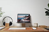 Wooden desk with a laptop perched on a stand, headphones, white wireless keyboard, and a white wireless mouse