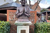 Statue of Mahatma Gandhi with his hands in prayer pose. Half a brick wall behind it and some hedges to either side. Plague reads: ‘May noble thoughts come to us from all directions” Mahatma Gandhi 1869–1948. Unveiled by Rt Hon The Lord Puttnam CBE. On the 15th October 2004.