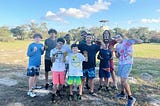 group of 8 campers and 2 activity leaders stand with their palms facing camera to show off their paint covered hands. all are looking at the camera and smiling. the rock climbing tower is seen in the background
