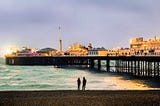 A man and a woman standing by the shore with a view of Brighton Pier, lit up for the evning