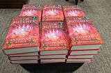 A table full of copies of The Olympian Affair by Jim Butcher on display at a bookstore. There are multiple stacks of the book. The books have a promotional $3 off price sticker.