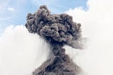 A gray cloud of smoke coming out of a volcano against a blue sky with white clouds.
