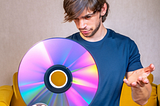 White man with shaggy hair and a bit of facial hair who is probably in his 20s is wearing an athletic blue t-shirt and holding a laser disc in one hand. His other hand is raised inquisitively and his stare is fixed on the laser disc with a facial expression that indicates he’s thinking something along the lines of, “What the fuck am supposed to do with this thing?” Image was AI generated.