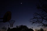 A crescent moon floats high above the last embers of a failing dusk. Silhouettes of trees, power lines, and a basketball hoop anchor the scene to the earth.