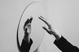 A black and white photo of a mirror on a white wall viewed from a slanted angle. A hand is extended towards the mirror, and its reflection is visible. The hand has white painted fingernails, a thin bracelet, and we can see the top of a black long-sleeve.