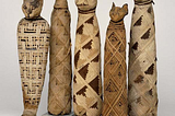 A row of five mummified cats in the British Museum. Each one is standing on a small square plinth, and essentially resembles a long tube wrapped in strips of (presumably) papyrus, in a variety of patterns, and with a carved (wooden?) cat’s head at the top