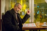 An older man sits in a cafe reading a newspaper.