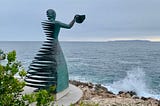 A bronze metal sculpture of a woman holding a handkerchief out as she faces the sea. On one side of the sculpture the metal is cut into horizontal ribbons. A wave breaks below on the rocky shore.