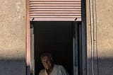 old man sitting in darkness, wearing a white shirt, looking out of his window.