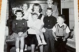Seven children in the 1950’s pose for a photo by a piano.